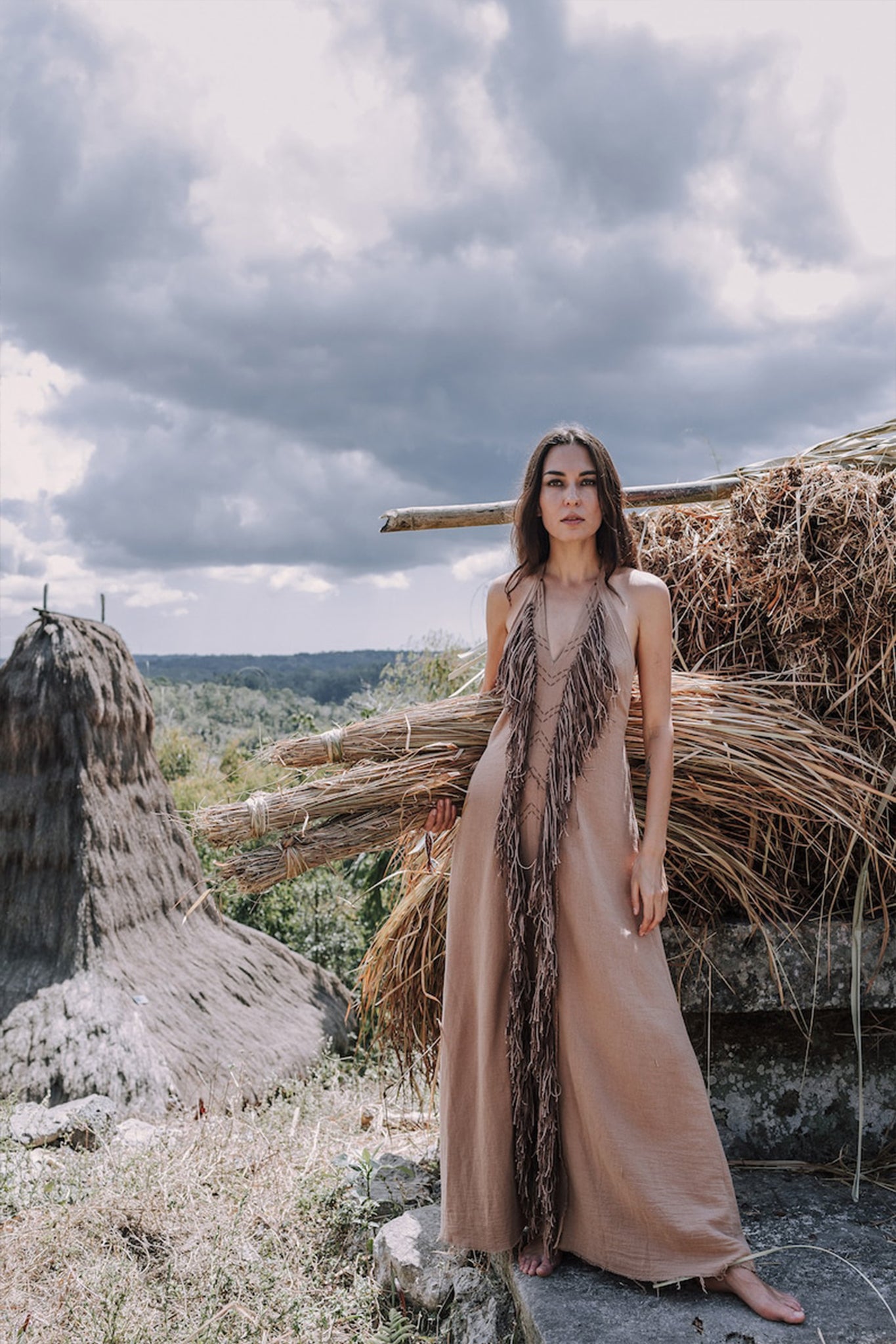 A woman stands barefoot in front of a rustic landscape, showcasing the Beige Organic Cotton Bohemian Tribal Maxi Dress with Hand Loomed Tassels by AYA Sacred Wear. The background features dramatic clouds and piles of dried grass.