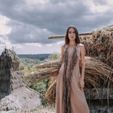 A woman stands barefoot in front of a rustic landscape, showcasing the Beige Organic Cotton Bohemian Tribal Maxi Dress with Hand Loomed Tassels by AYA Sacred Wear. The background features dramatic clouds and piles of dried grass.