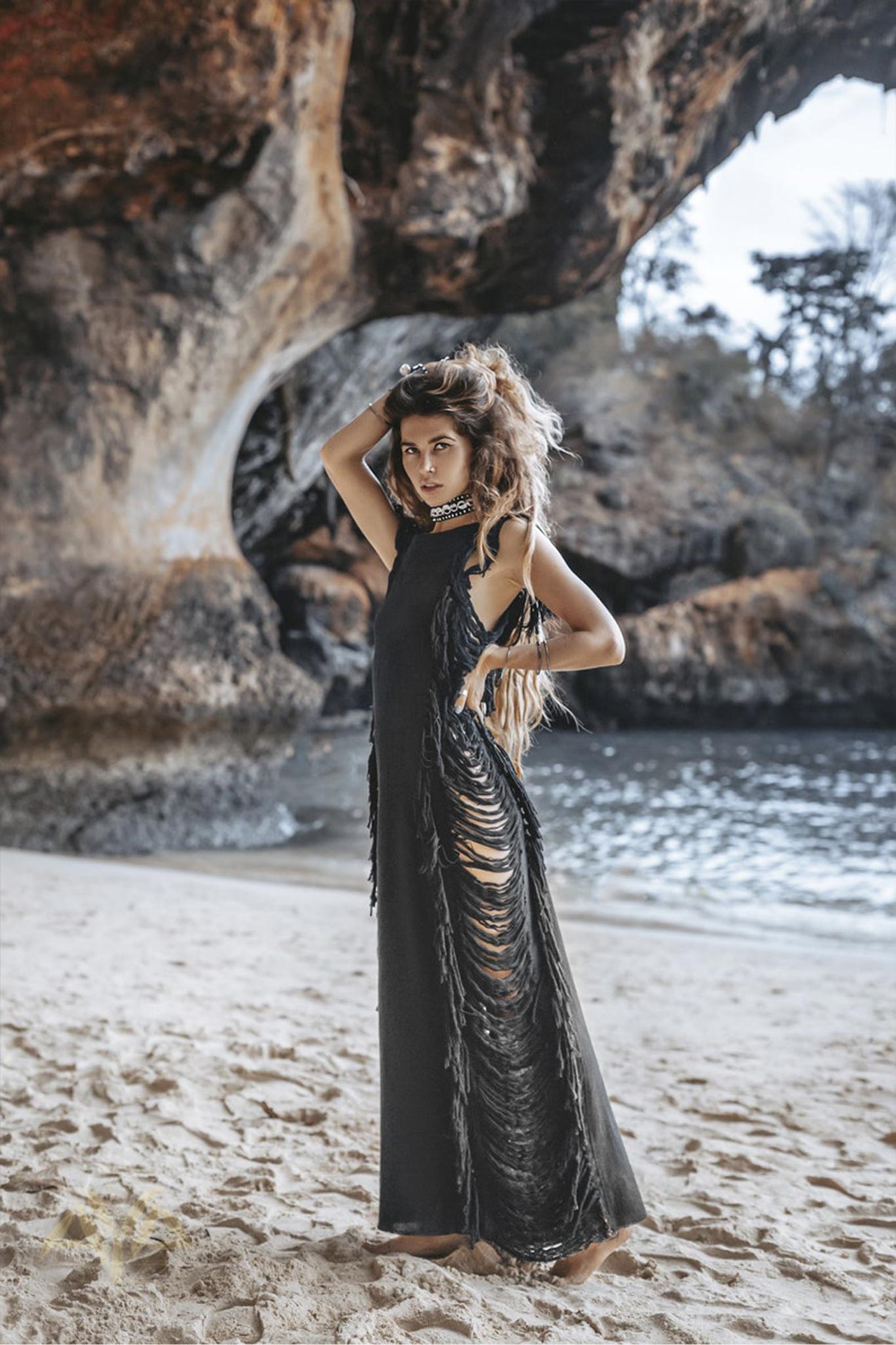 A woman in an elegant Black Boho Dress by AYA Sacred Wear stands on a sandy beach under a rocky archway. Wearing the Bohemian style dress featuring ladder sides, she poses with her hand on her head, gazing at the camera. The ocean and trees are visible in the background.