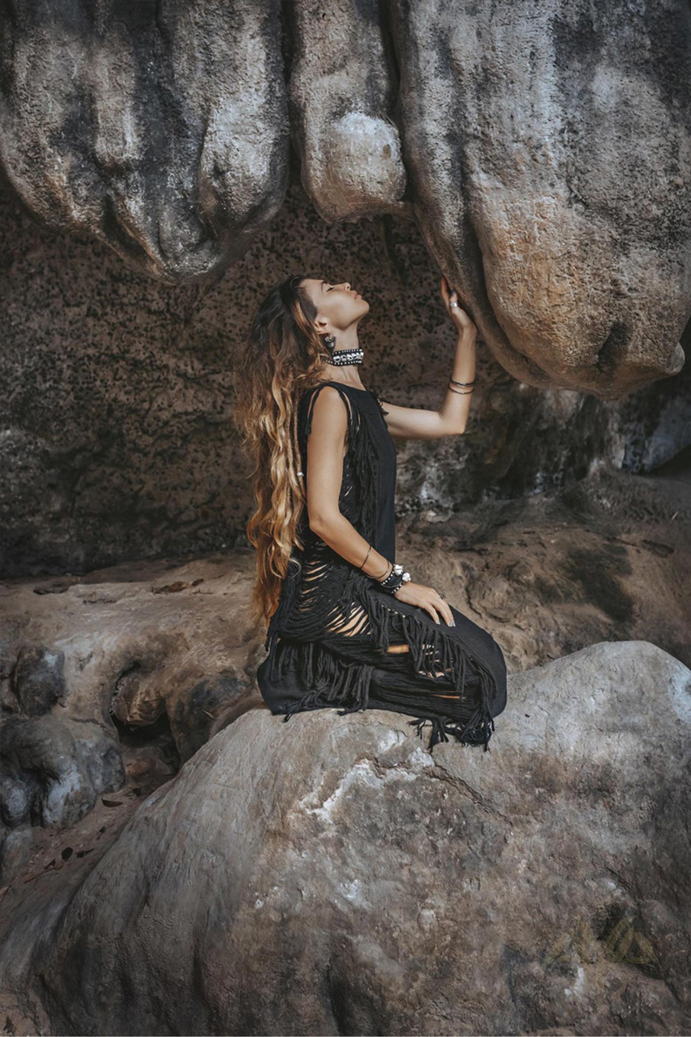 A person with long hair kneels on a rock, gazing up at towering stone formations. They are wearing an elegant Ladder Sides Dress from AYA Sacred Wear, adorned with bracelets and necklaces. The black attire enhances the serene juxtaposition of rugged natural rocks and intricate craftsmanship.