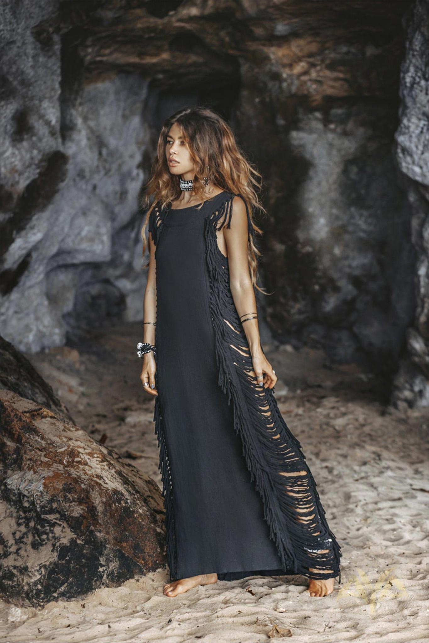 A woman with long hair stands barefoot on sandy ground against a rocky cave backdrop. She wears the AYA Sacred Wear Black Boho Dress, featuring ladder sides and fringe detailing, along with various bracelets for a bohemian look.