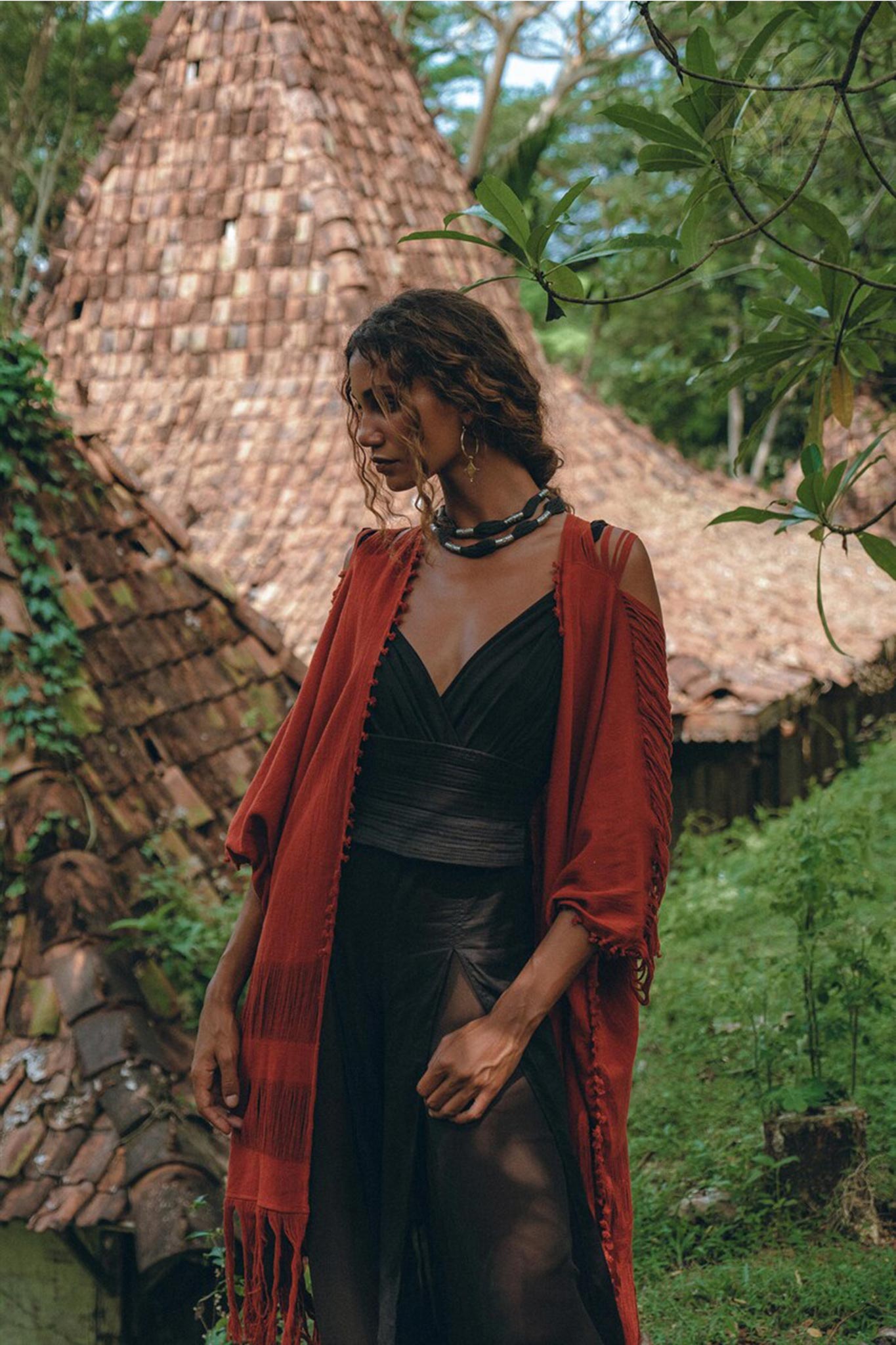 A woman wearing a black boho jumpsuit, a handmade cold shoulder maxi creation by AYA Sacred Wear, stands in front of a thatched-roof cottage surrounded by lush greenery. She is draped in a red shawl and looks down, with trees providing a serene and natural backdrop.