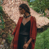 A woman wearing a black boho jumpsuit, a handmade cold shoulder maxi creation by AYA Sacred Wear, stands in front of a thatched-roof cottage surrounded by lush greenery. She is draped in a red shawl and looks down, with trees providing a serene and natural backdrop.
