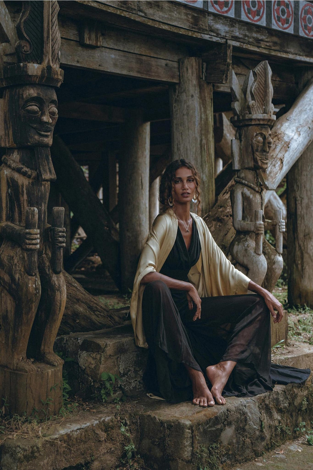 A woman wearing a Black Boho Jumpsuit by AYA Sacred Wear, featuring handmade cold shoulder details, sits barefoot on a stone step near intricately carved wooden statues under a rustic wooden structure. The setting is enveloped in an ancient, tribal vibe highlighted by natural earth tones and textures.