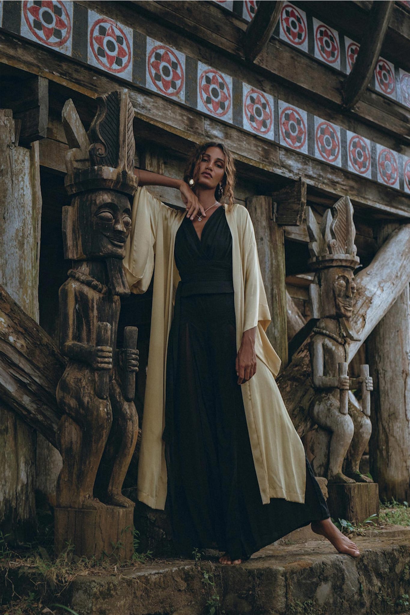 A woman wearing a Black Boho Jumpsuit from AYA Sacred Wear, featuring a handmade cold shoulder maxi design, stands leaning against a wooden sculpture. She poses with one arm resting on her head, surrounded by tribal carvings and a rustic wooden structure with intricate red and white patterns.