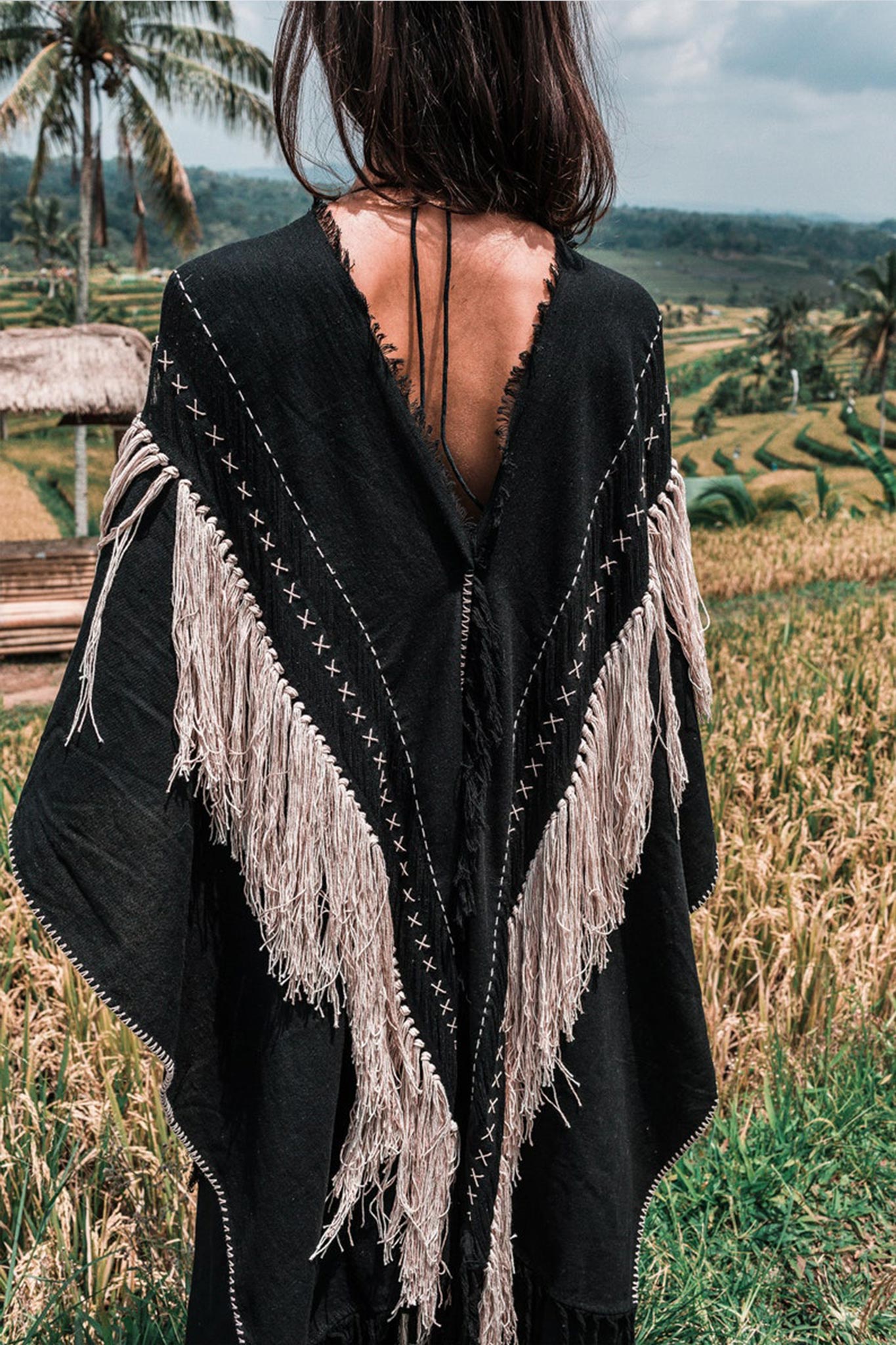 A woman with long dark hair stands in a field, enveloped in the AYA Sacred Wear Black Boho Poncho, adorned with hand stitching and hand loomed tassels. The background features terraced greenery and palm trees beneath a cloudy sky.