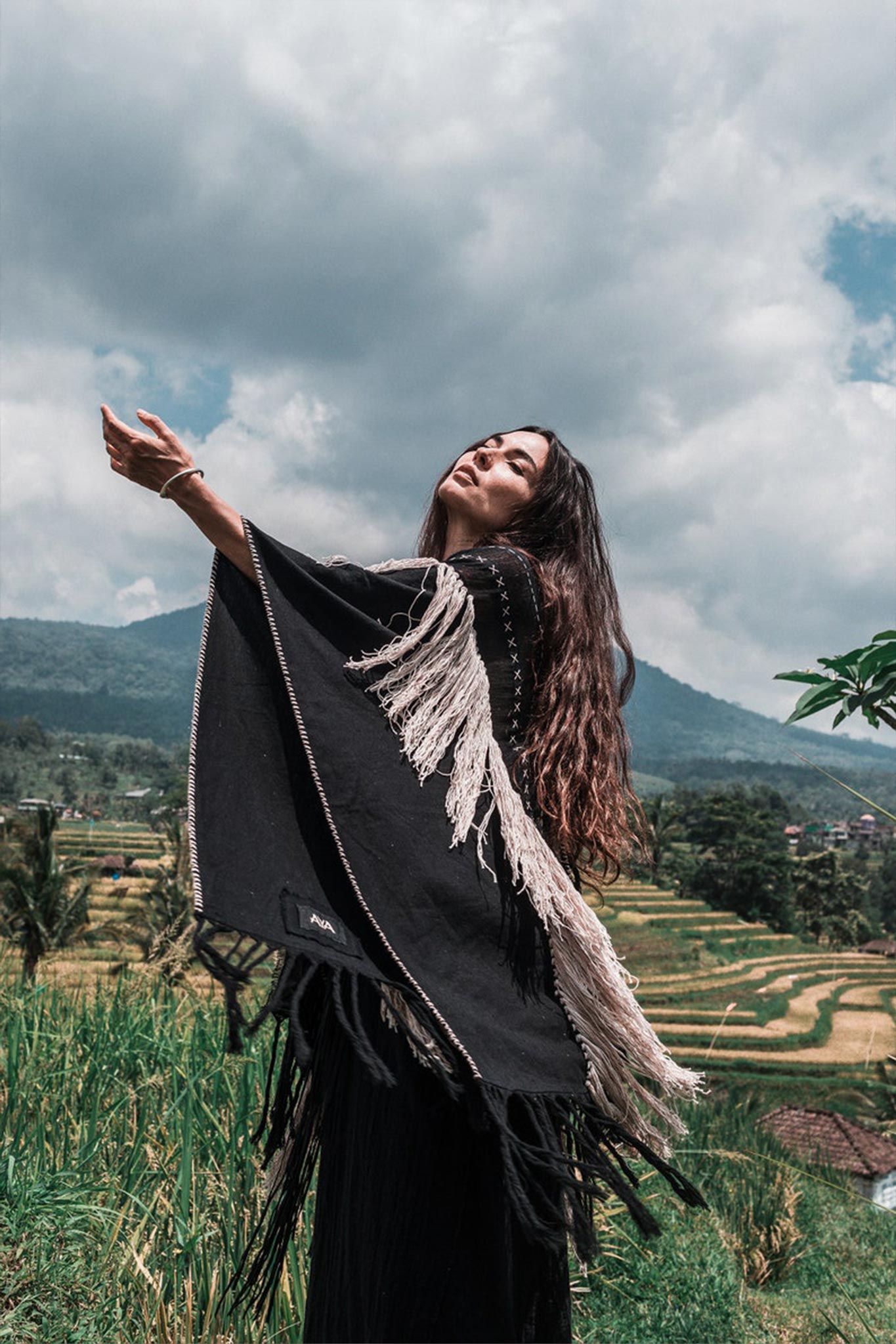 In a lush green field, a woman with long hair stands adorned in an AYA Sacred Wear Black Boho Poncho. This hand-stitched mantel, featuring hand-loomed tassels and made from organic cotton, complements the majestic backdrop of mountains and a cloudy sky as she slightly raises her arms, embodying a sense of freedom and harmony with nature.