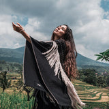 In a lush green field, a woman with long hair stands adorned in an AYA Sacred Wear Black Boho Poncho. This hand-stitched mantel, featuring hand-loomed tassels and made from organic cotton, complements the majestic backdrop of mountains and a cloudy sky as she slightly raises her arms, embodying a sense of freedom and harmony with nature.