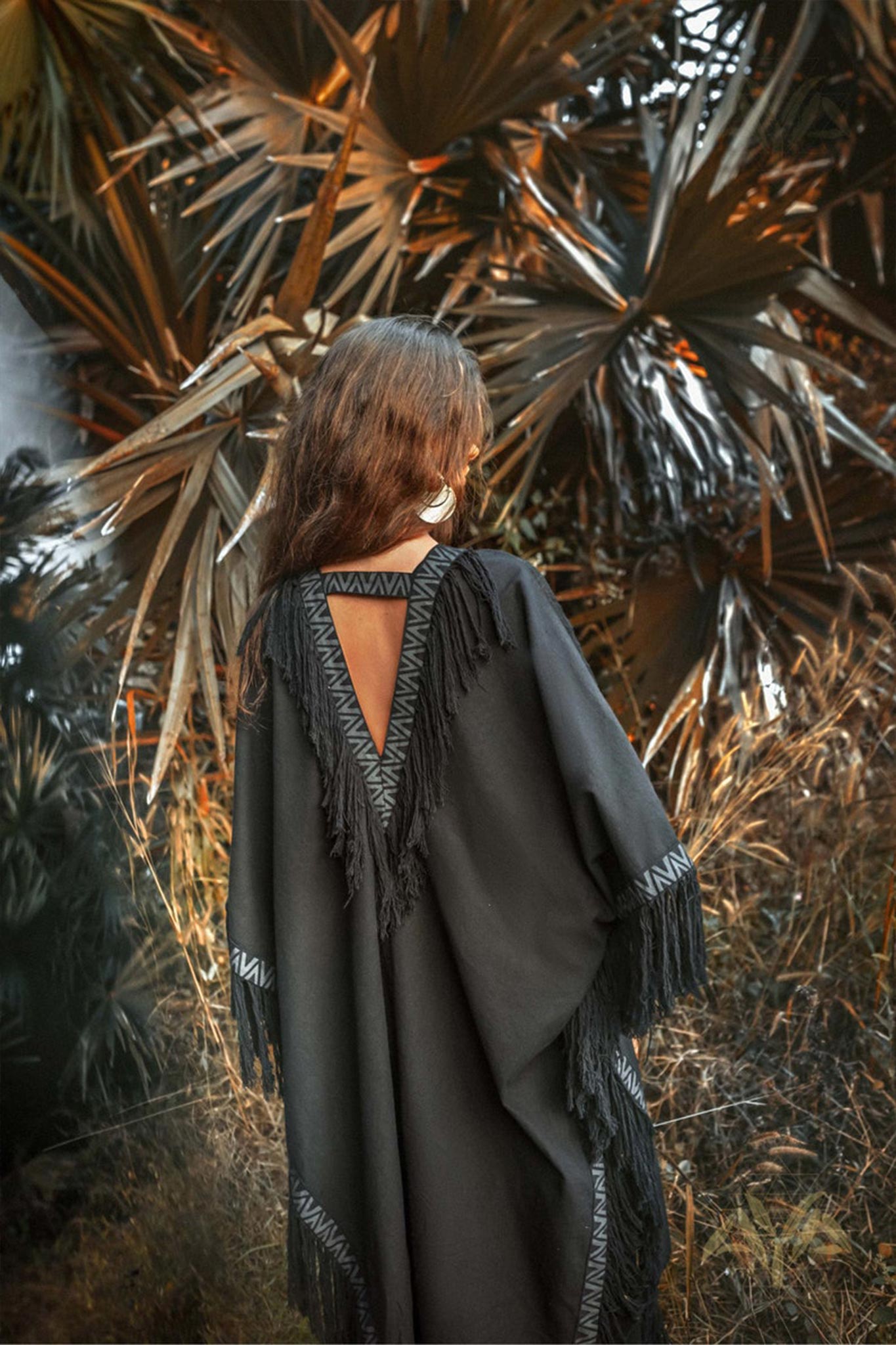 Amidst large, dark palm leaves, a woman with long hair wears the Black Boho Poncho by AYA Sacred Wear, made from organic cotton. The plus size unisex poncho features a fringed, triangular open-back design that complements her relaxed pose as she faces away from the camera, perfectly merging with the natural surroundings.