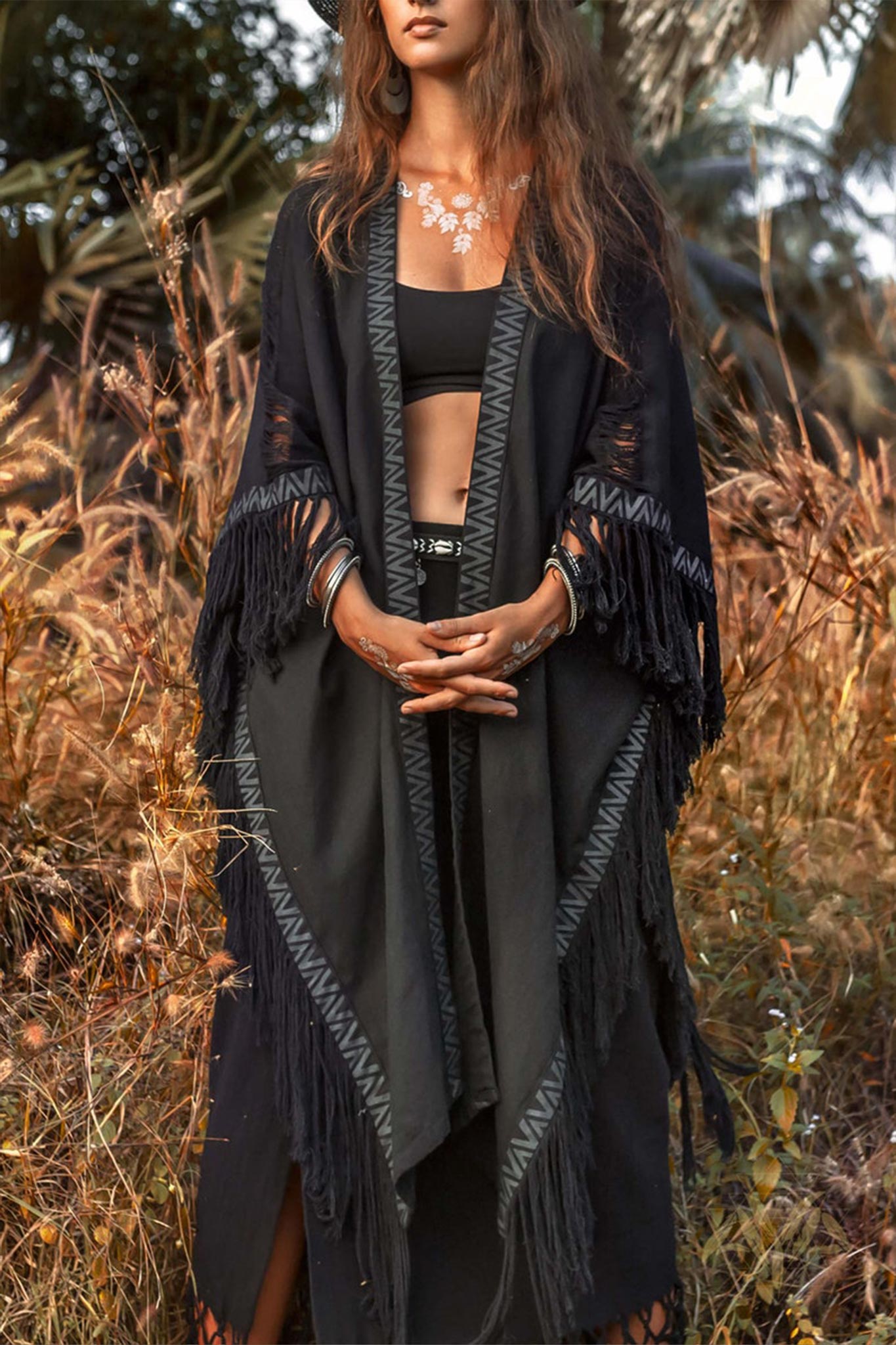 A person stands in tall grass wearing the Black Boho Poncho from AYA Sacred Wear over a black top and skirt. With long hair, they are adorned with a floral necklace and various jewelry, their arms crossed. The hand-loomed poncho enhances the earthy vibe against a backdrop of trees.