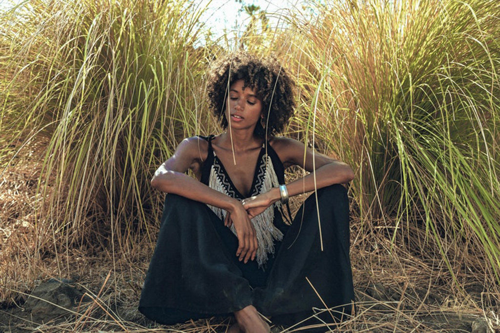 Amidst the tall grasses, a person with curly hair peacefully sits wearing a Black Boho Tunic Top with Tassels and Hand Embroidery by AYA Sacred Wear. Their eyes are closed, and their hands rest on their knees in a serene outdoor setting.