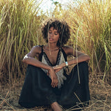 Amidst the tall grasses, a person with curly hair peacefully sits wearing a Black Boho Tunic Top with Tassels and Hand Embroidery by AYA Sacred Wear. Their eyes are closed, and their hands rest on their knees in a serene outdoor setting.