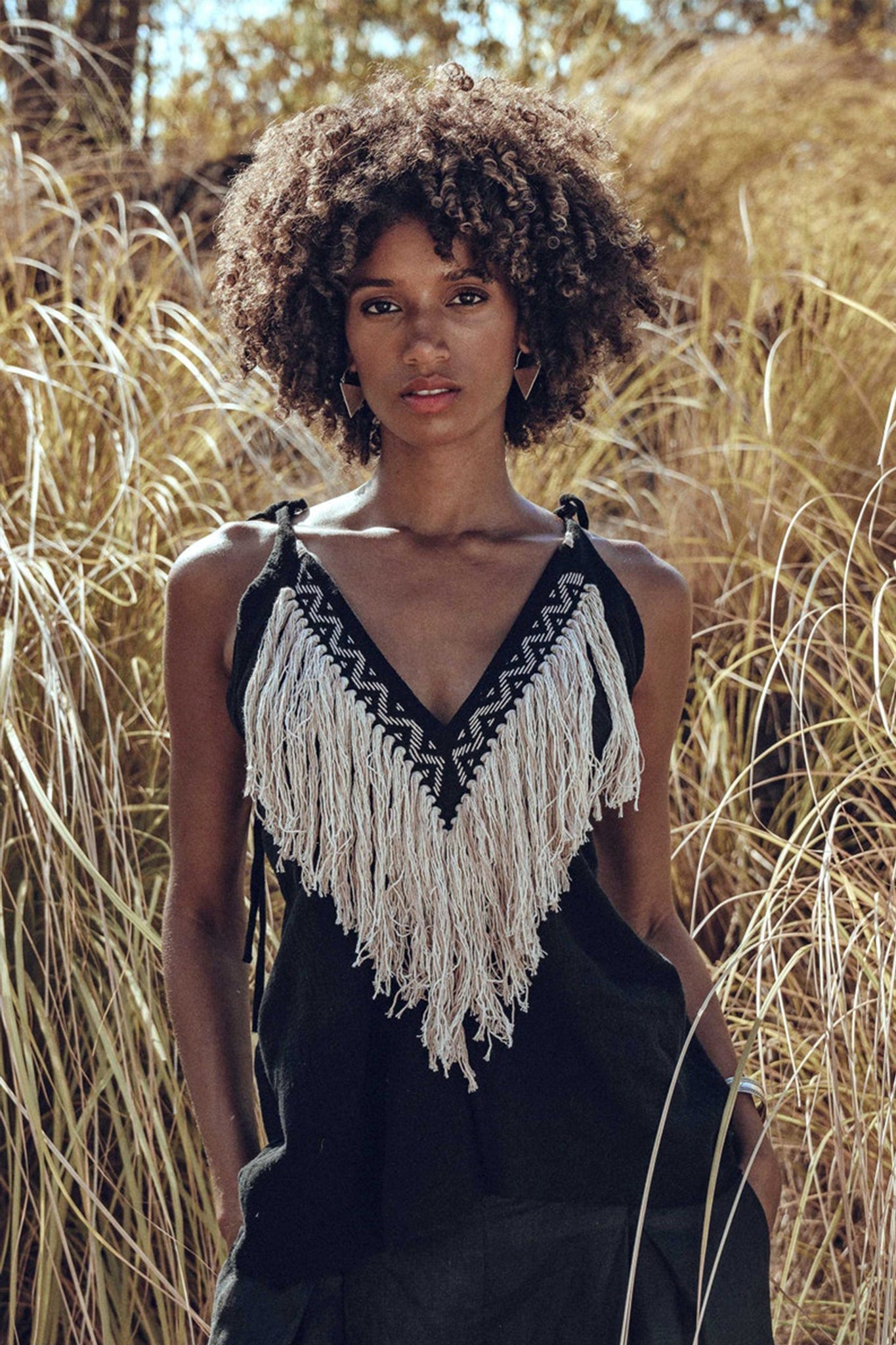 A person with curly hair stands outdoors amidst tall grasses, wearing the Black Boho Tunic Top by AYA Sacred Wear, featuring tassels and hand embroidery. The sunlight highlights the natural scenery while beautifully complementing the hand-loomed cotton fabric's texture.