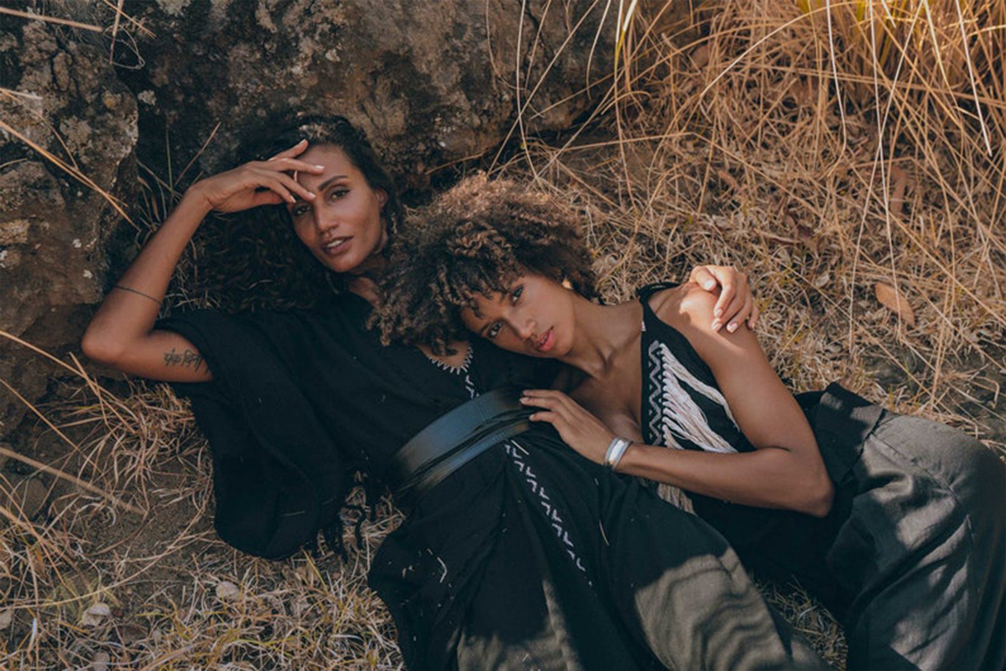 Two individuals dressed in AYA Sacred Wear's Black Boho Tunic Top with Tassels and Hand Embroidery lie on the ground in a dry, grassy area. One rests their head on the other's shoulder as they both gaze toward the camera, surrounded by rocks and dried vegetation.