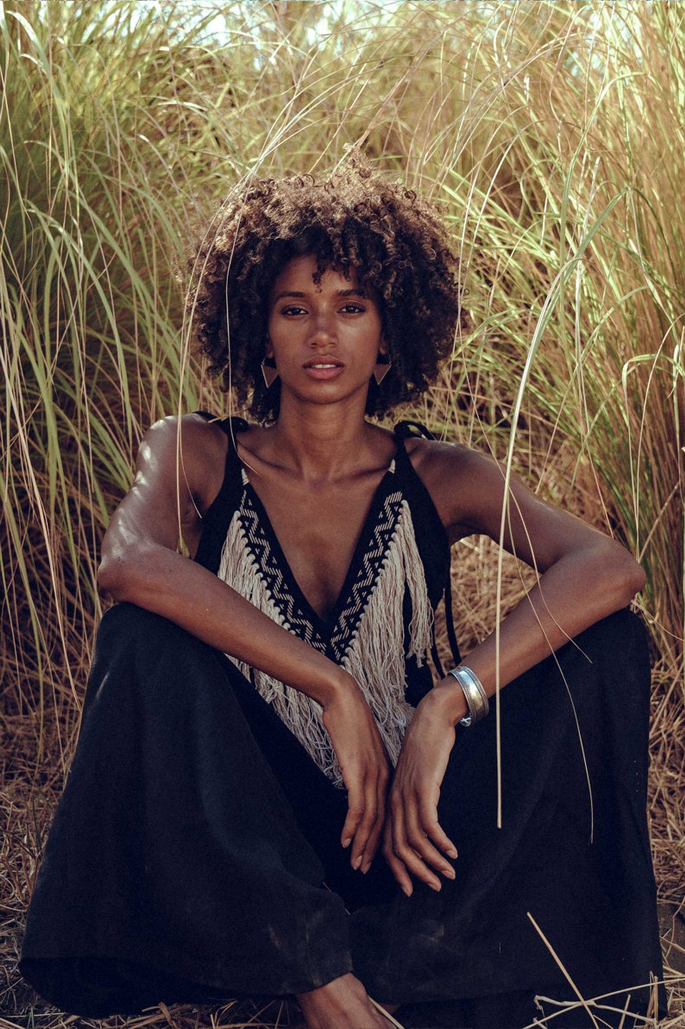 Amidst the tall grass, a person with curly hair sits serenely, dressed in a black outfit featuring intricate front detailing and a silver bracelet. The sunlight highlights their calm expression, while the Black Boho Tunic Top with Tassels and Hand Embroidery by AYA Sacred Wear, made from hand-loomed cotton, adds an earthy touch to the scene.