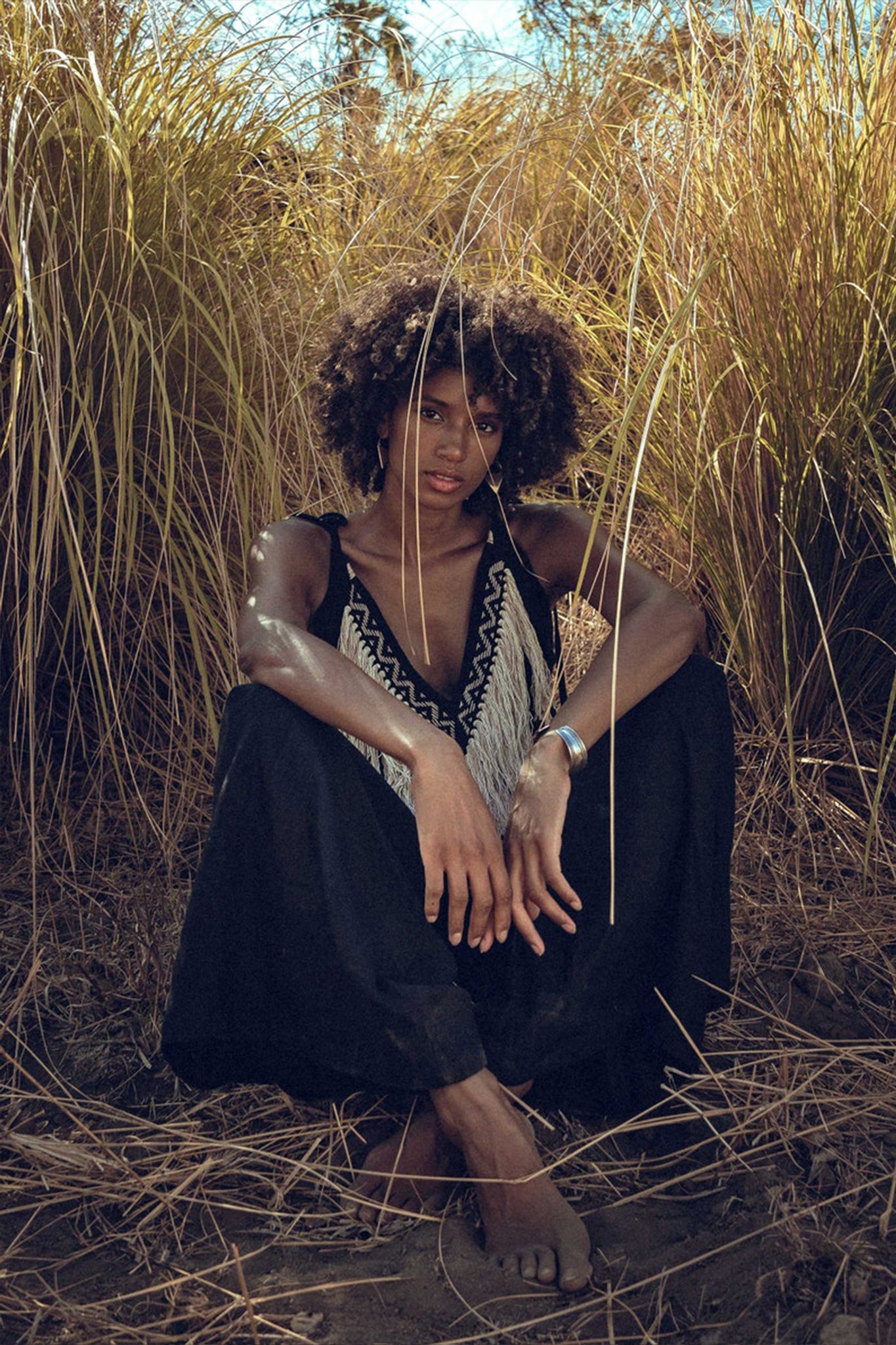 A person with curly hair sits among tall, dry grass, sporting a sleeveless Black Boho Tunic Top with Tassels and Hand Embroidery from AYA Sacred Wear and black pants. They have a thoughtful expression and a silver bracelet on their right wrist, surrounded by a natural and warm setting.