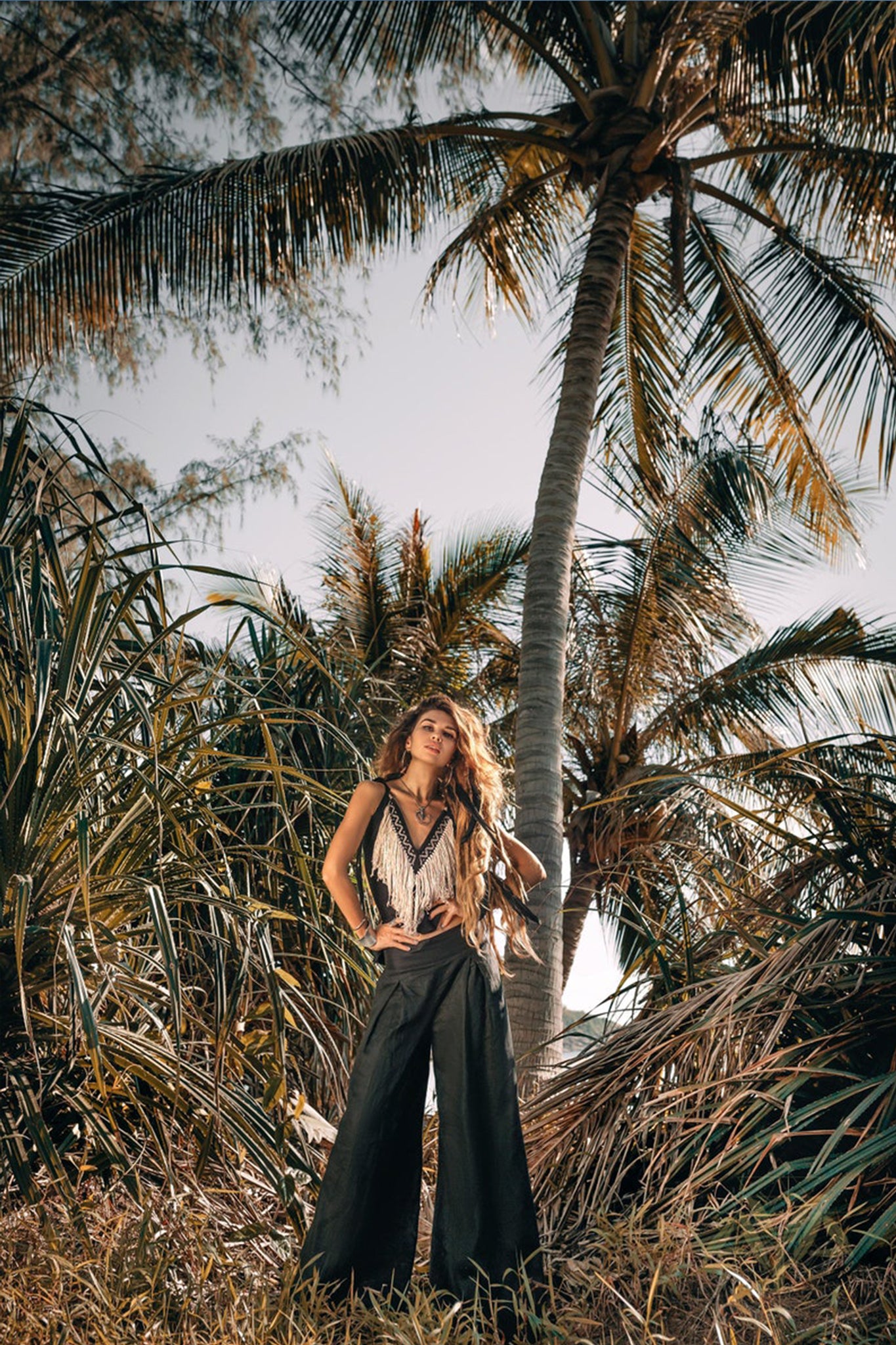 Standing confidently among tall palm trees and lush greenery, a person wears the Black Boho Tunic Top with Tassels and Hand Embroidery by AYA Sacred Wear, paired with wide-leg pants crafted from hand-loomed cotton. The sunlight highlights their long flowing hair, creating a serene tropical atmosphere that celebrates sustainable fashion.