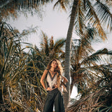 Standing confidently among tall palm trees and lush greenery, a person wears the Black Boho Tunic Top with Tassels and Hand Embroidery by AYA Sacred Wear, paired with wide-leg pants crafted from hand-loomed cotton. The sunlight highlights their long flowing hair, creating a serene tropical atmosphere that celebrates sustainable fashion.