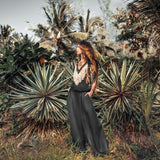 A woman with long hair stands in a tropical setting, surrounded by lush green foliage and spiky plants. She wears the Black Boho Tunic Top with Tassels and Hand Embroidery from AYA Sacred Wear and wide pants made from hand-loomed cotton, looking off into the distance under a clear sky.