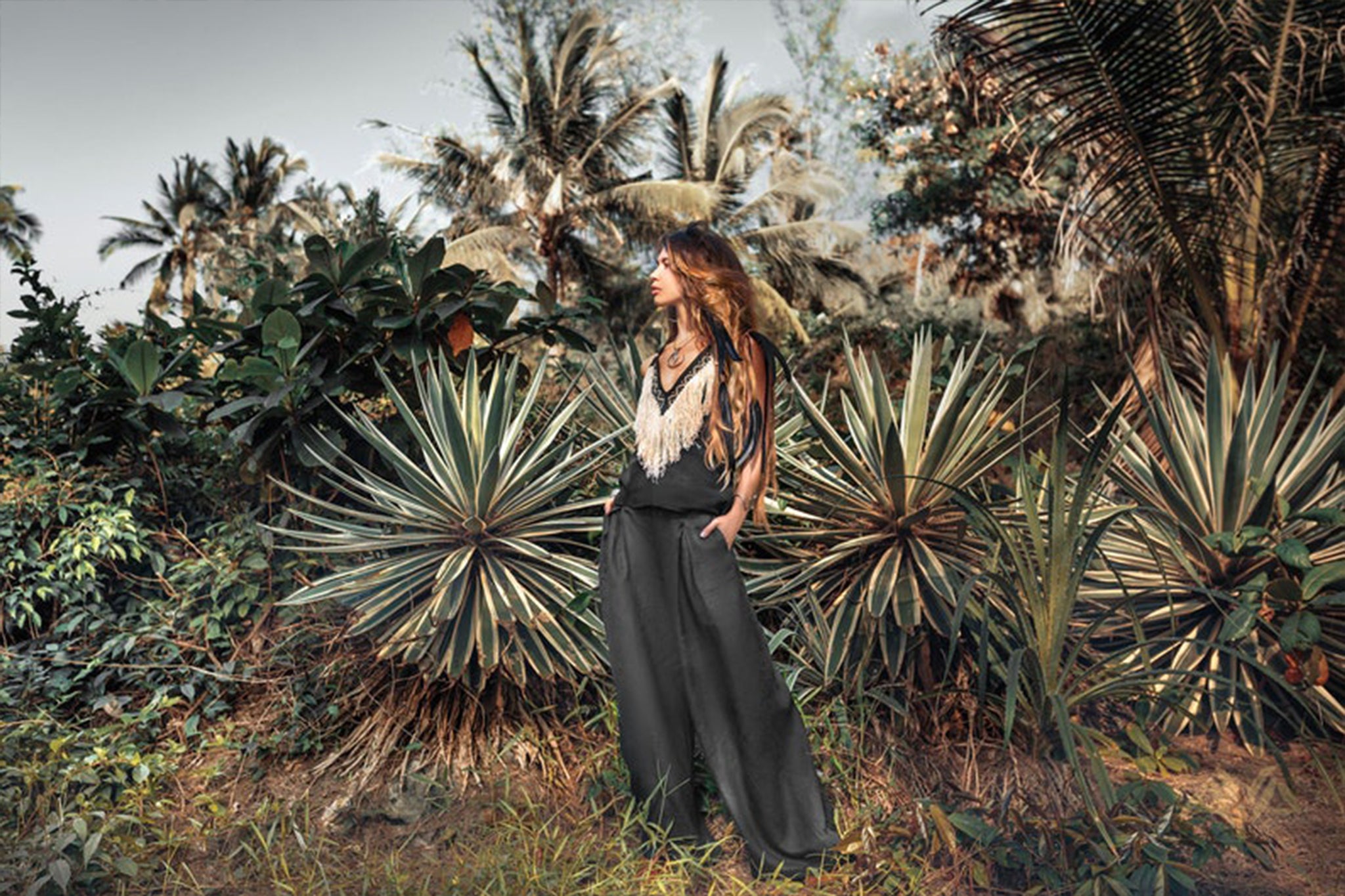 A woman with long hair stands in a tropical setting, surrounded by lush green foliage and spiky plants. She wears the Black Boho Tunic Top with Tassels and Hand Embroidery from AYA Sacred Wear and wide pants made from hand-loomed cotton, looking off into the distance under a clear sky.