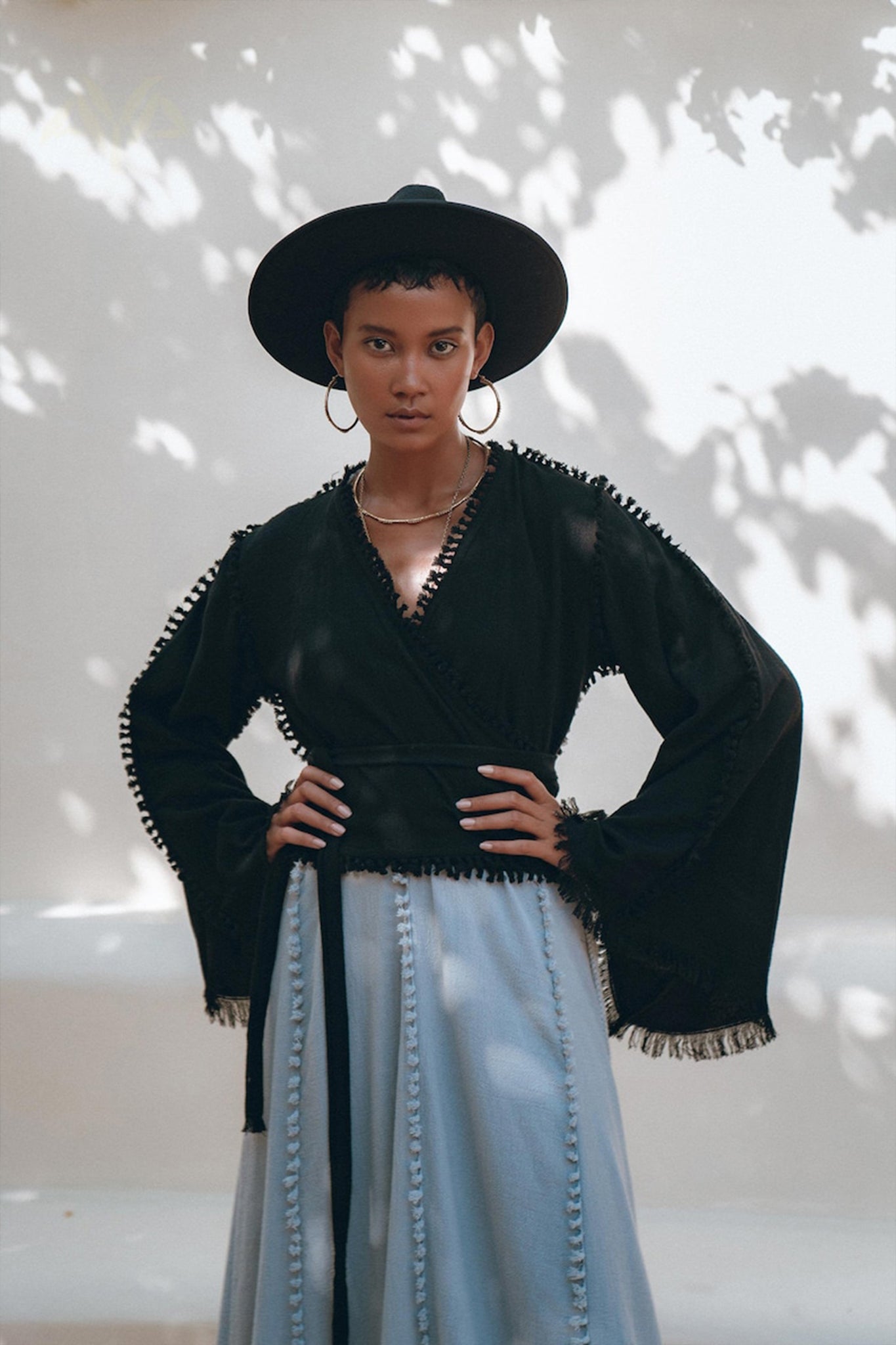 A fashionista poses confidently outdoors, wearing a black wide-brimmed hat and the Black Boho Wrap Top with wide sleeves by AYA Sacred Wear. Paired with a gray skirt featuring decorative details, her ensemble shines in the dappled sunlight, while tree shadow patterns on the wall behind enhance the scene.