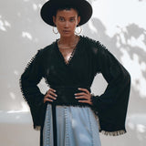 A fashionista poses confidently outdoors, wearing a black wide-brimmed hat and the Black Boho Wrap Top with wide sleeves by AYA Sacred Wear. Paired with a gray skirt featuring decorative details, her ensemble shines in the dappled sunlight, while tree shadow patterns on the wall behind enhance the scene.