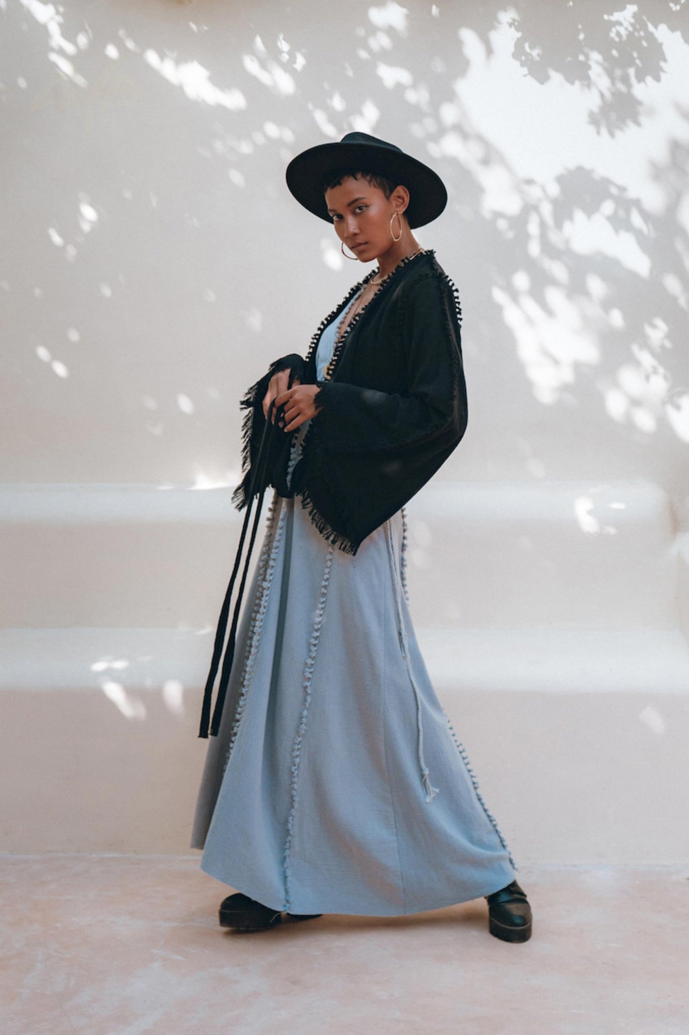 A person poses outdoors in a stylish outfit, featuring a wide-brimmed black hat, the Black Boho Wrap Top from AYA Sacred Wear with its durable fringe details and wide sleeves, and a long light blue dress. The background is softly lit with dappled shadows from nearby foliage.