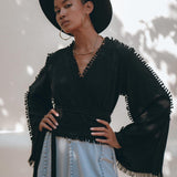 A person wearing a wide-brimmed black hat, gold hoop earrings, and the Black Boho Wrap Top from AYA Sacred Wear poses confidently. They have their hands on their hips and wear a light blue skirt. Shadowed leaves create a pattern on the light background.