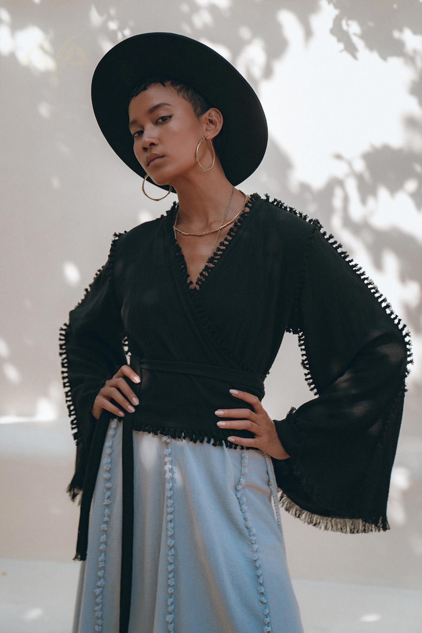 A person wearing a wide-brimmed black hat, gold hoop earrings, and the Black Boho Wrap Top from AYA Sacred Wear poses confidently. They have their hands on their hips and wear a light blue skirt. Shadowed leaves create a pattern on the light background.