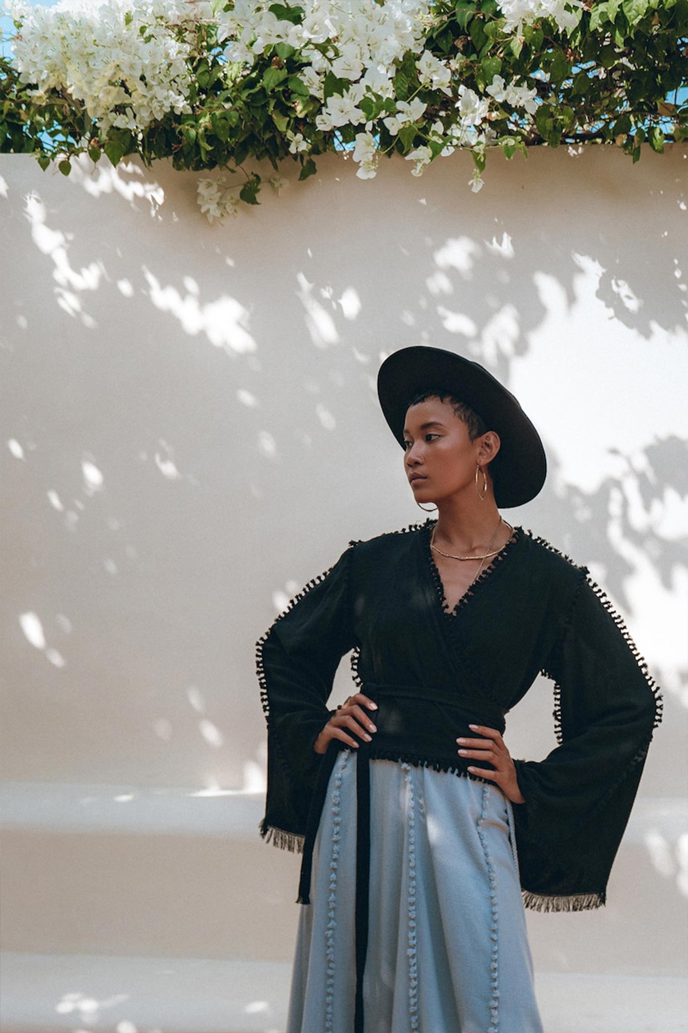 A person stands confidently against a light-colored wall with dappled sunlight. They wear a wide-brimmed black hat, the Black Boho Wrap Top from AYA Sacred Wear, featuring organic cotton fabric and fringed sleeves, paired with light blue trousers. White flowers hang above, adding a natural touch.