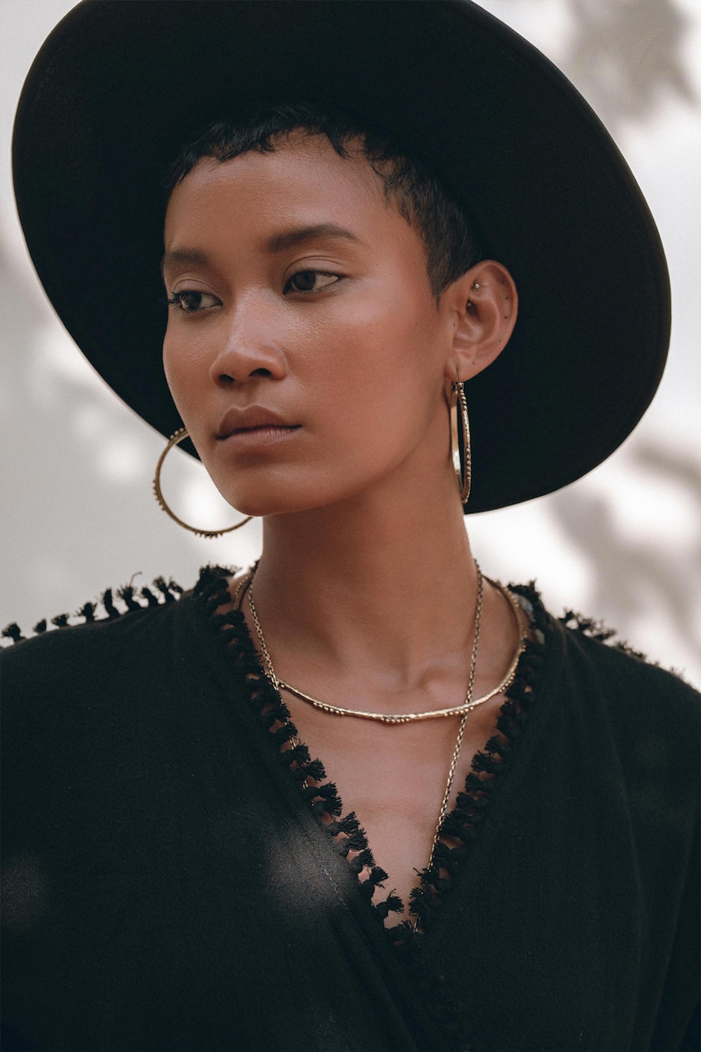 A person is elegantly dressed in an AYA Sacred Wear Black Boho Wrap Top featuring wide sleeves and fringe details. Crafted from organic cotton, this high-quality garment exudes both style and durability. They are accessorized with a large black hat, hoop earrings, and layered necklaces, while their calm and thoughtful expression stands out against a softly blurred background with dappled light.