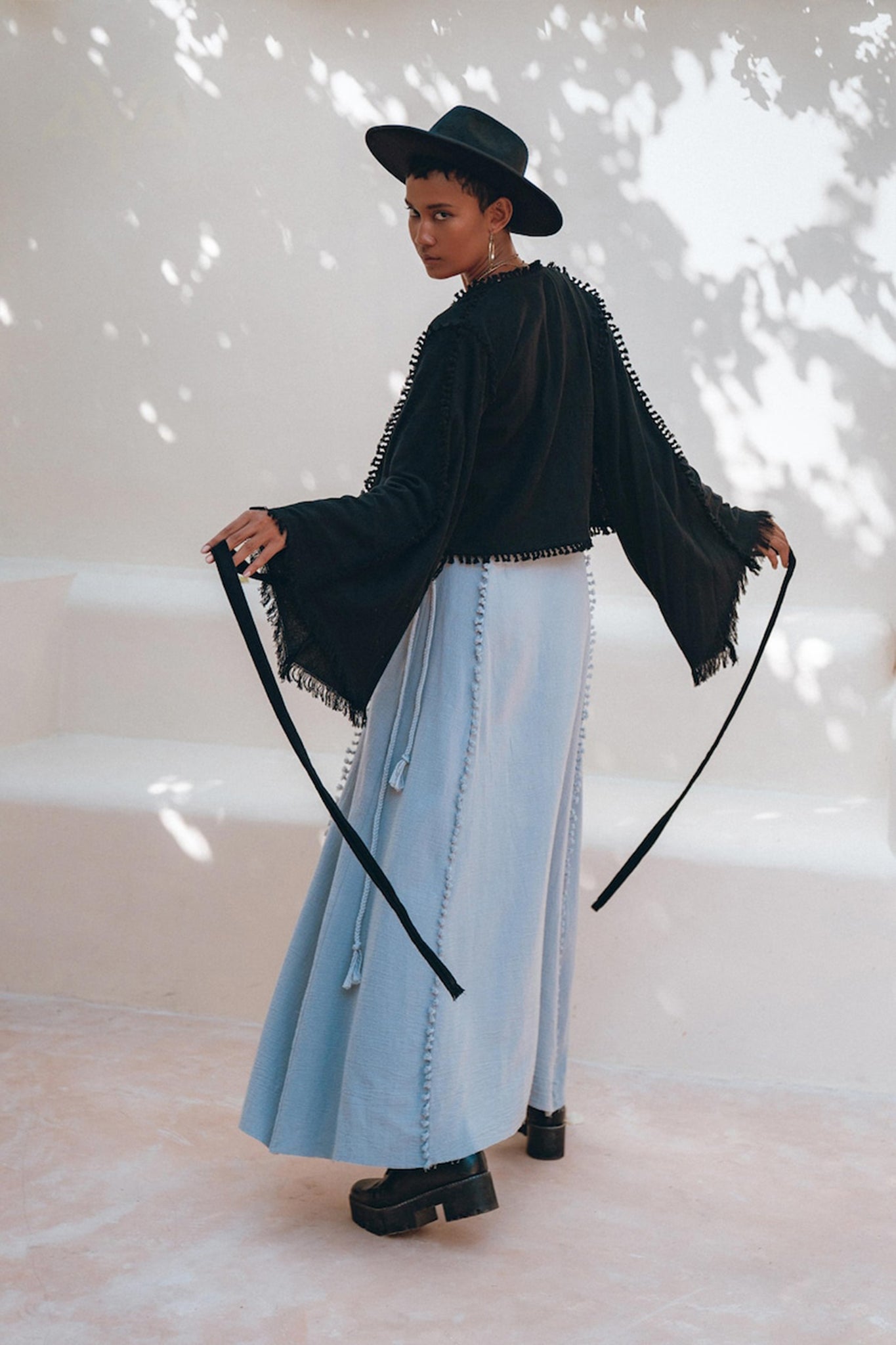 An individual wearing the AYA Sacred Wear's Black Boho Wrap Top, featuring wide sleeves and fringe details, paired with a light blue organic cotton skirt, stands facing backward while tying a knot. The softly lit background creates a captivating shadow effect, allowing their handmade design to shine through the ensemble.