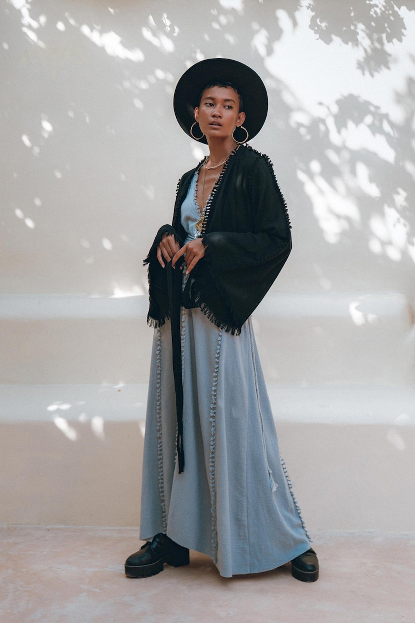 A person stands confidently wearing a black wide-brimmed hat, large hoop earrings, and a black fringed jacket. They pair this ensemble with the Black Boho Wrap Top from AYA Sacred Wear, crafted from organic cotton with decorative trim. The outfit is completed by a long light blue dress that adds an ethereal touch to the look. They pose against a neutral backdrop with soft shadows for an effortlessly chic appearance.