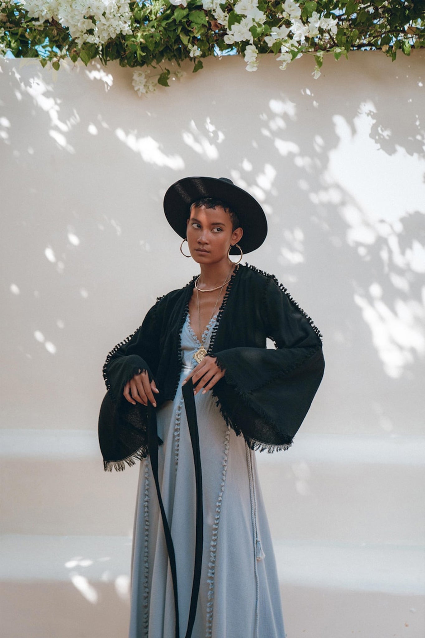 A woman wearing the Black Boho Wrap Top from AYA Sacred Wear, complemented by a wide-brimmed hat and a long light dress, stands against a sunlit wall with dappled shadows. The ensemble features a handcrafted design, with white flowers hanging above to create an artful scene.