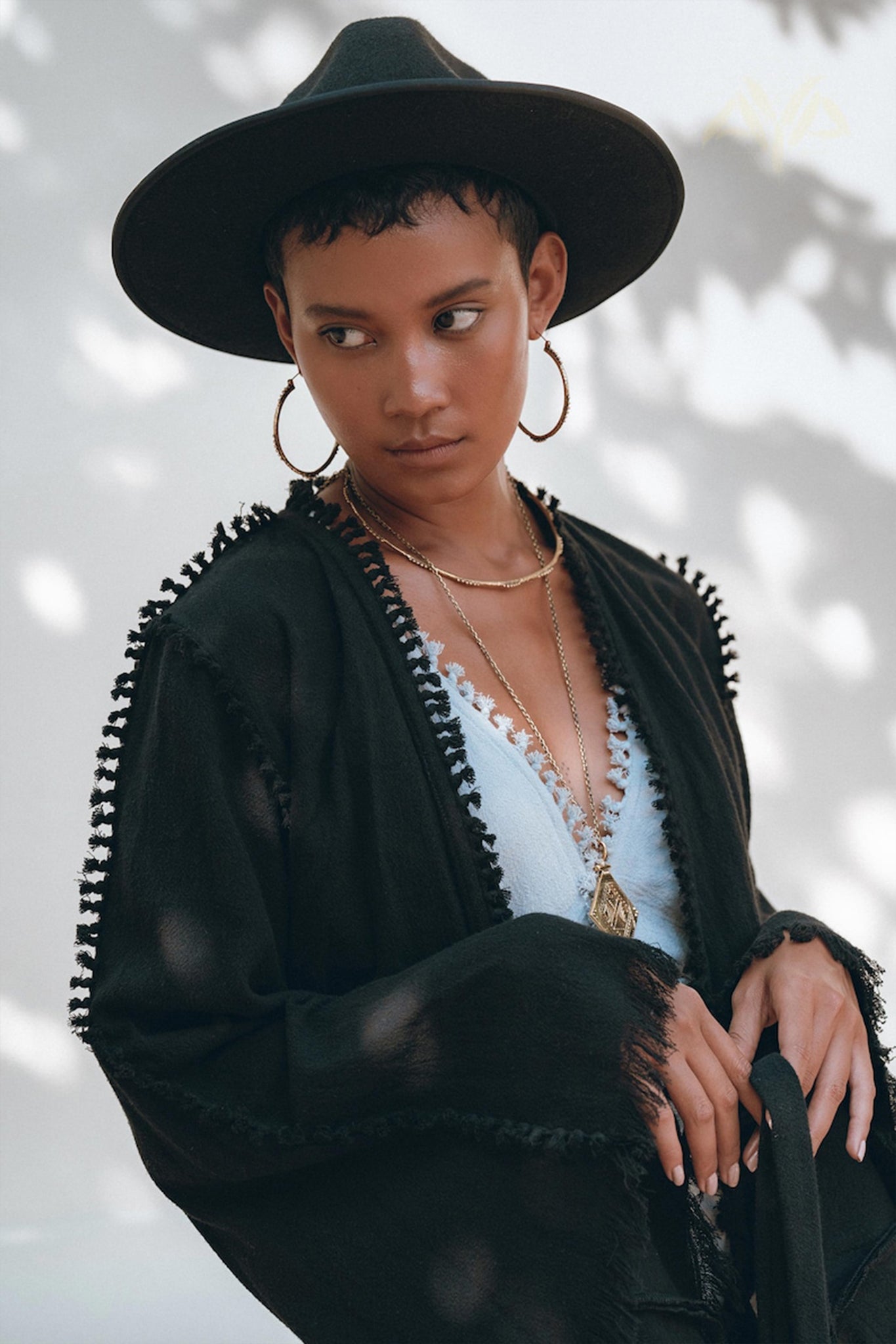 A person wearing a wide-brimmed hat and the Black Boho Wrap Top by AYA Sacred Wear stands against a softly blurred light background with abstract shadows. Accessorized with hoop earrings and layered necklaces, their gaze is directed to the side. This outfit features the handmade design elements of the Wide Sleeve Organic Top Women, adding a unique touch.