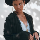 A person wearing a wide-brimmed hat and the Black Boho Wrap Top by AYA Sacred Wear stands against a softly blurred light background with abstract shadows. Accessorized with hoop earrings and layered necklaces, their gaze is directed to the side. This outfit features the handmade design elements of the Wide Sleeve Organic Top Women, adding a unique touch.