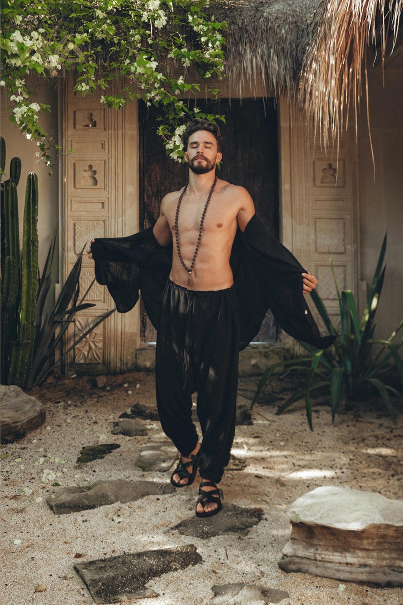 A bearded man stands outdoors in front of a decorative stone wall, wearing AYA Sacred Wear's Black Colour Handwoven Cotton Pants for Men. He pairs the pants with sandals and an open black shirt that reveals his chest adorned with a beaded necklace, surrounded by lush greenery and rocky ground.