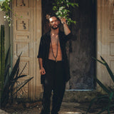 A man with dark hair and a beard stands in front of a rustic wooden door, wearing an open black shirt and AYA Sacred Wear's Black Colour Handwoven Cotton Pants for Men, paired with sandals. He holds a leafy vine above him, surrounded by a lush, green garden.