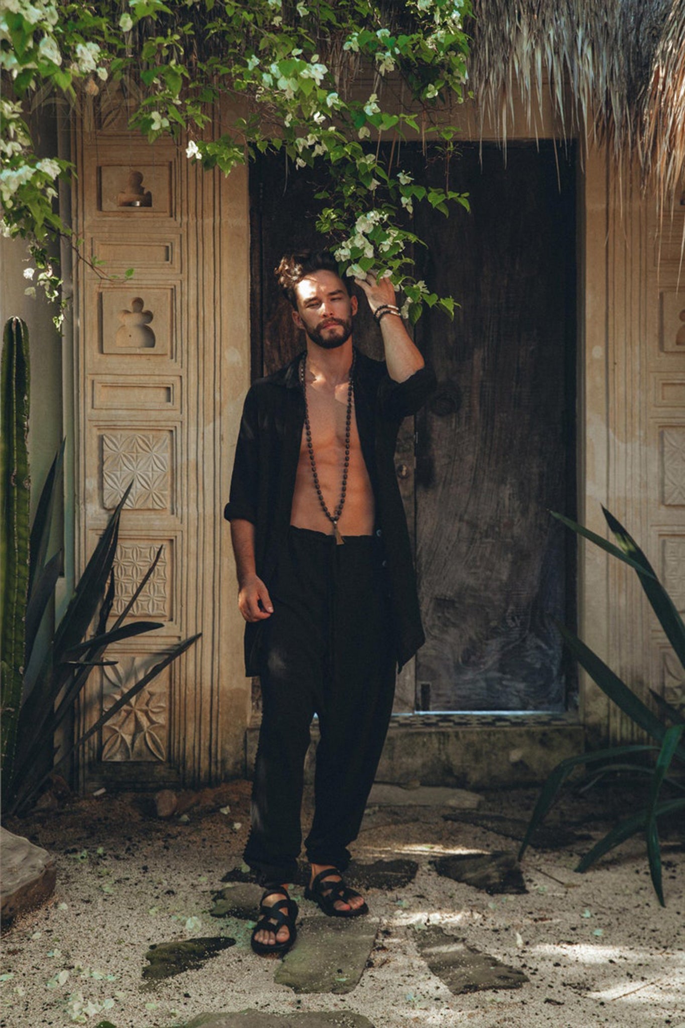 A man with dark hair and a beard stands in front of a rustic wooden door, wearing an open black shirt and AYA Sacred Wear's Black Colour Handwoven Cotton Pants for Men, paired with sandals. He holds a leafy vine above him, surrounded by a lush, green garden.