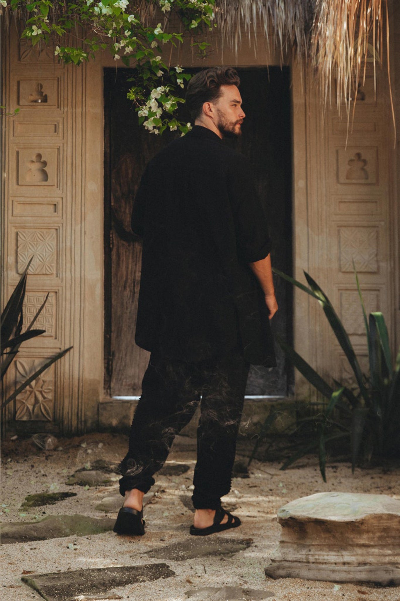 A person dressed in AYA Sacred Wear's Black Colour Handwoven Cotton Pants for Men stands on a sandy pathway before an ornate wooden door, enveloped by lush plants and greenery, enhancing the tranquil and natural ambiance of the scene.