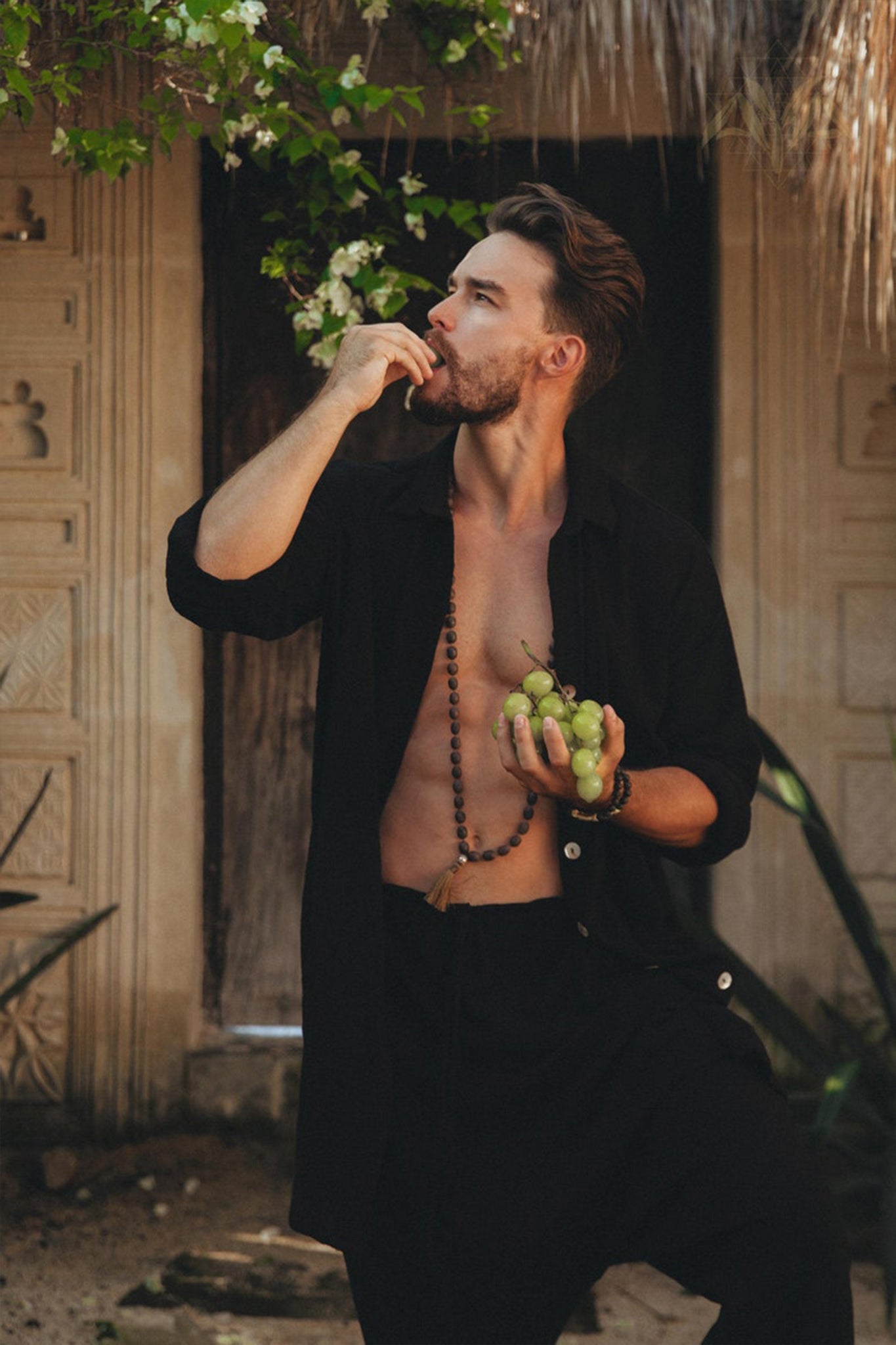 A man stands outdoors, enjoying some green grapes while wearing a partially open black shirt and a pair of AYA Sacred Wear's Black Colour Handwoven Cotton Pants for Men, enhancing his relaxed vibe. The background includes a rustic wooden door and plants with sunlight gently filtering through.