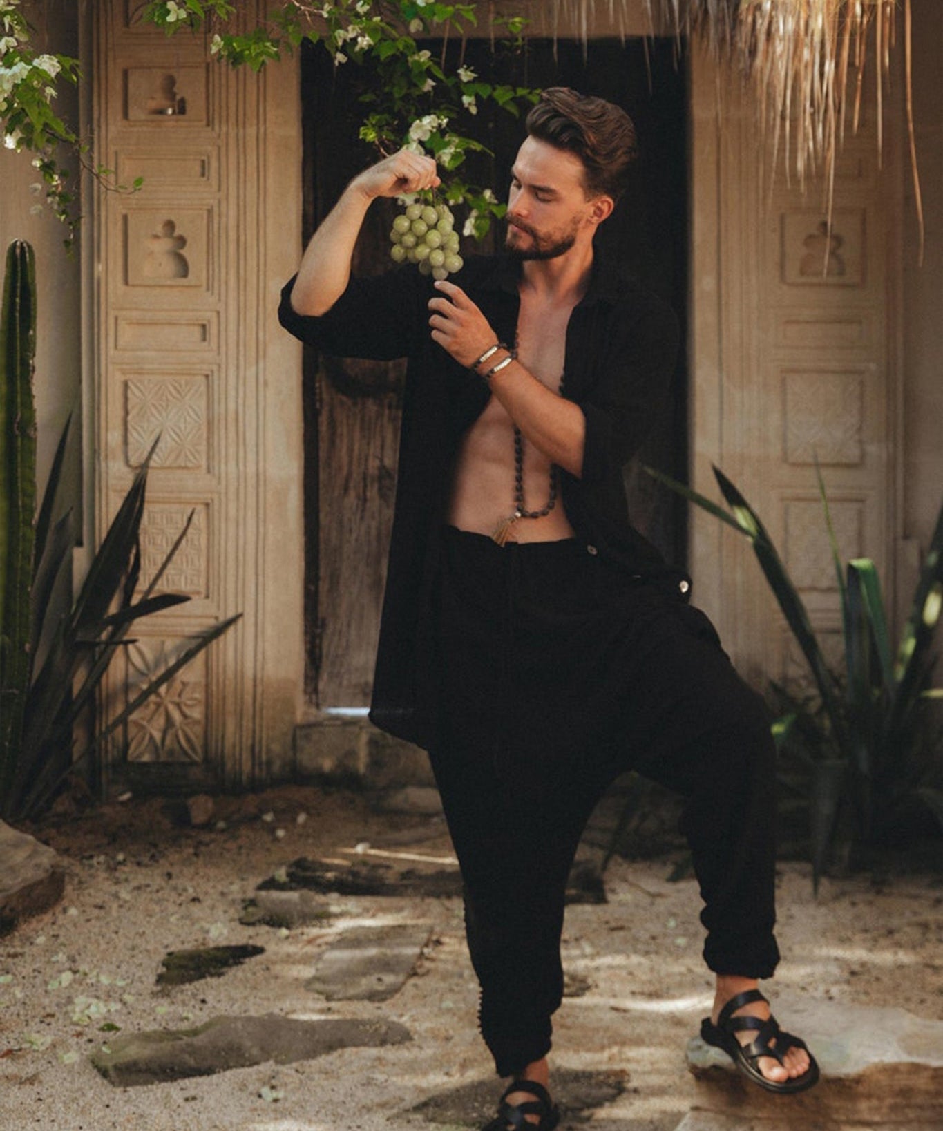 Outdoors in a tropical setting with lush greenery and an ornate, carved doorway in the background, a bearded man is holding a bunch of grapes. He is dressed in an open black shirt paired with Black Colour Handwoven Cotton Pants for Men by AYA Sacred Wear.