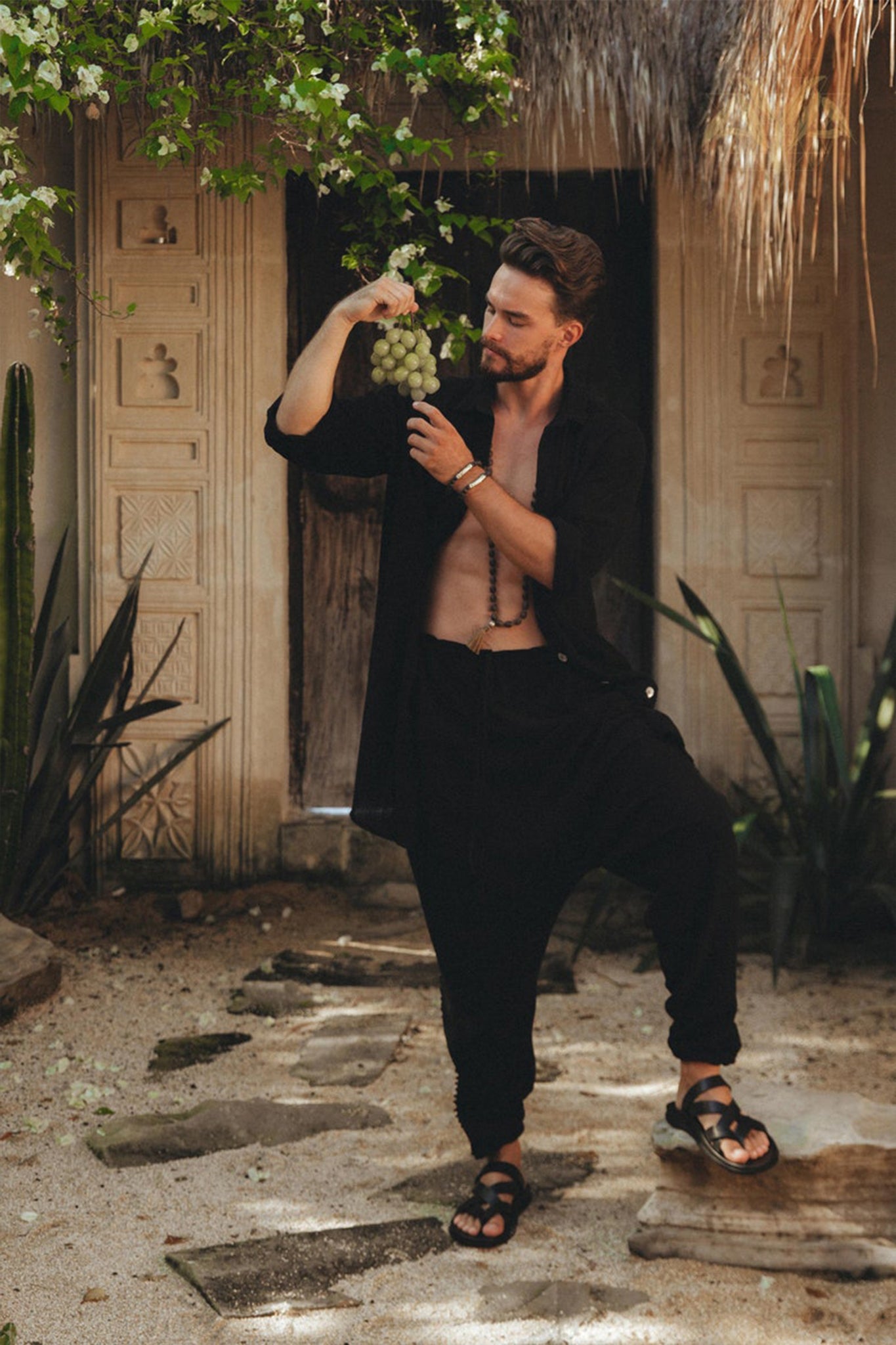 Outdoors in a tropical setting with lush greenery and an ornate, carved doorway in the background, a bearded man is holding a bunch of grapes. He is dressed in an open black shirt paired with Black Colour Handwoven Cotton Pants for Men by AYA Sacred Wear.