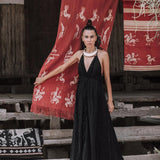 A woman wearing the AYA Sacred Wear Black Cotton Boho Maxi Dress with Hand Loomed Tassels stands in front of a red woven textile featuring white patterns, set outdoors. Her hair is styled up, and she accessorizes with a white necklace. The background showcases wooden structures and additional textiles.