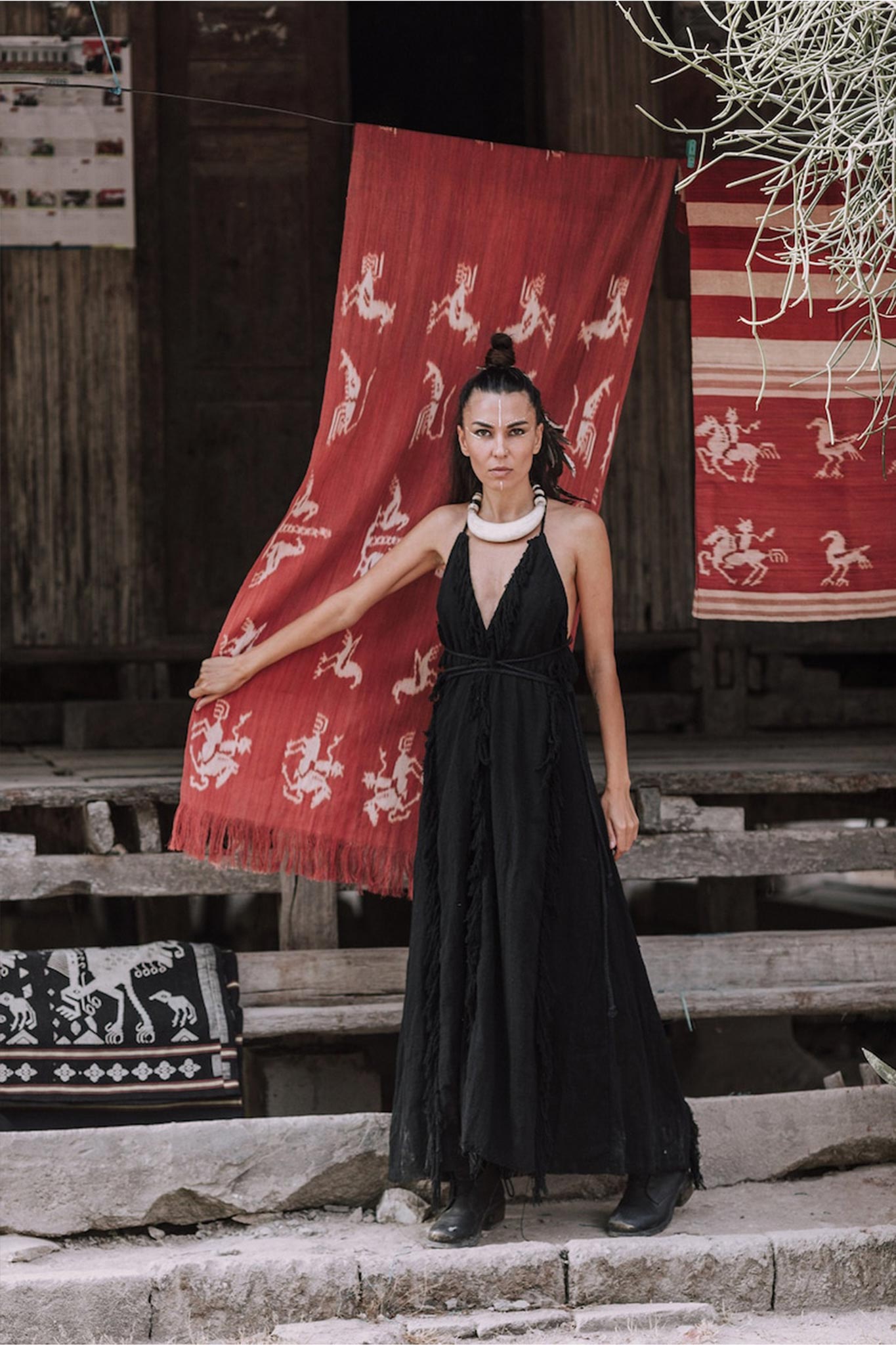 A woman wearing the AYA Sacred Wear Black Cotton Boho Maxi Dress with Hand Loomed Tassels stands in front of a red woven textile featuring white patterns, set outdoors. Her hair is styled up, and she accessorizes with a white necklace. The background showcases wooden structures and additional textiles.