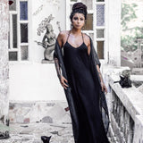 A woman in a Black Cotton Boho Wedding Slip Dress by AYA Sacred Wear stands on a stone terrace beside a black cat. Behind her is a wall adorned with stained glass windows and a statue. Her long dark hair is styled in an updo, she wears a necklace, and the cat sits at her feet.