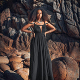 A woman stands confidently amidst rugged terrain, dressed in an elegant Black Greek Goddess Prom Boho Evening Dress from AYA Sacred Wear. Her hands rest on her hips, and her voluminous hair is beautifully highlighted by the warm, natural light against a backdrop of large stones and jagged rocks.
