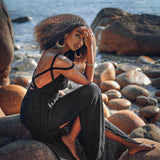 A woman in a flowing Black Greek Goddess Prom Boho Evening Dress by AYA Sacred Wear sits on rocky terrain by the sea. Her curly hair is complemented by large hoop earrings and bracelets, while the sun glistens on her skin as she gazes at the camera.