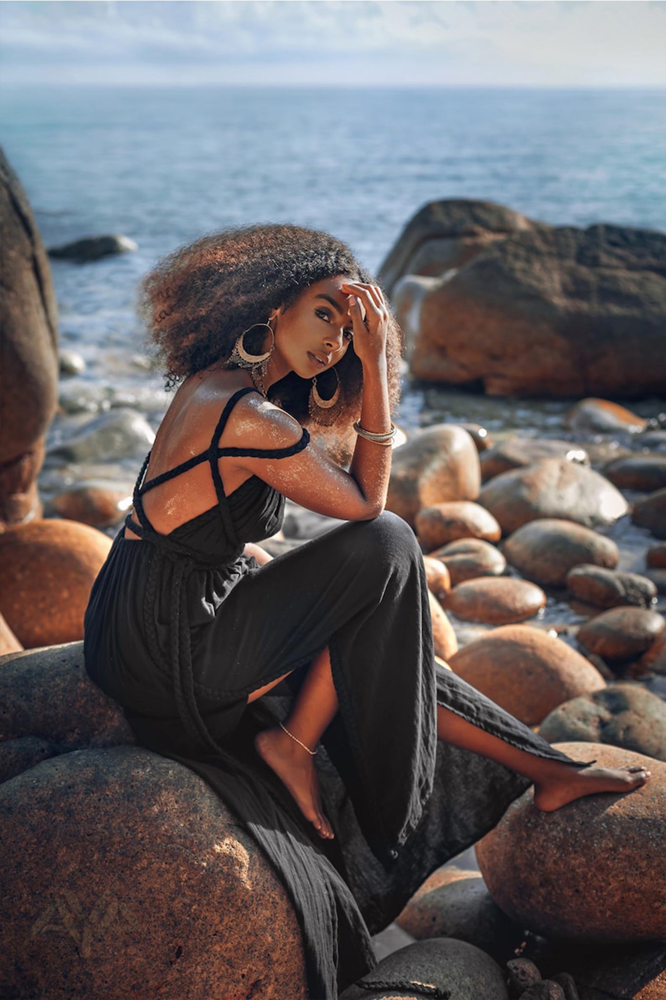 A woman in a flowing Black Greek Goddess Prom Boho Evening Dress by AYA Sacred Wear sits on rocky terrain by the sea. Her curly hair is complemented by large hoop earrings and bracelets, while the sun glistens on her skin as she gazes at the camera.