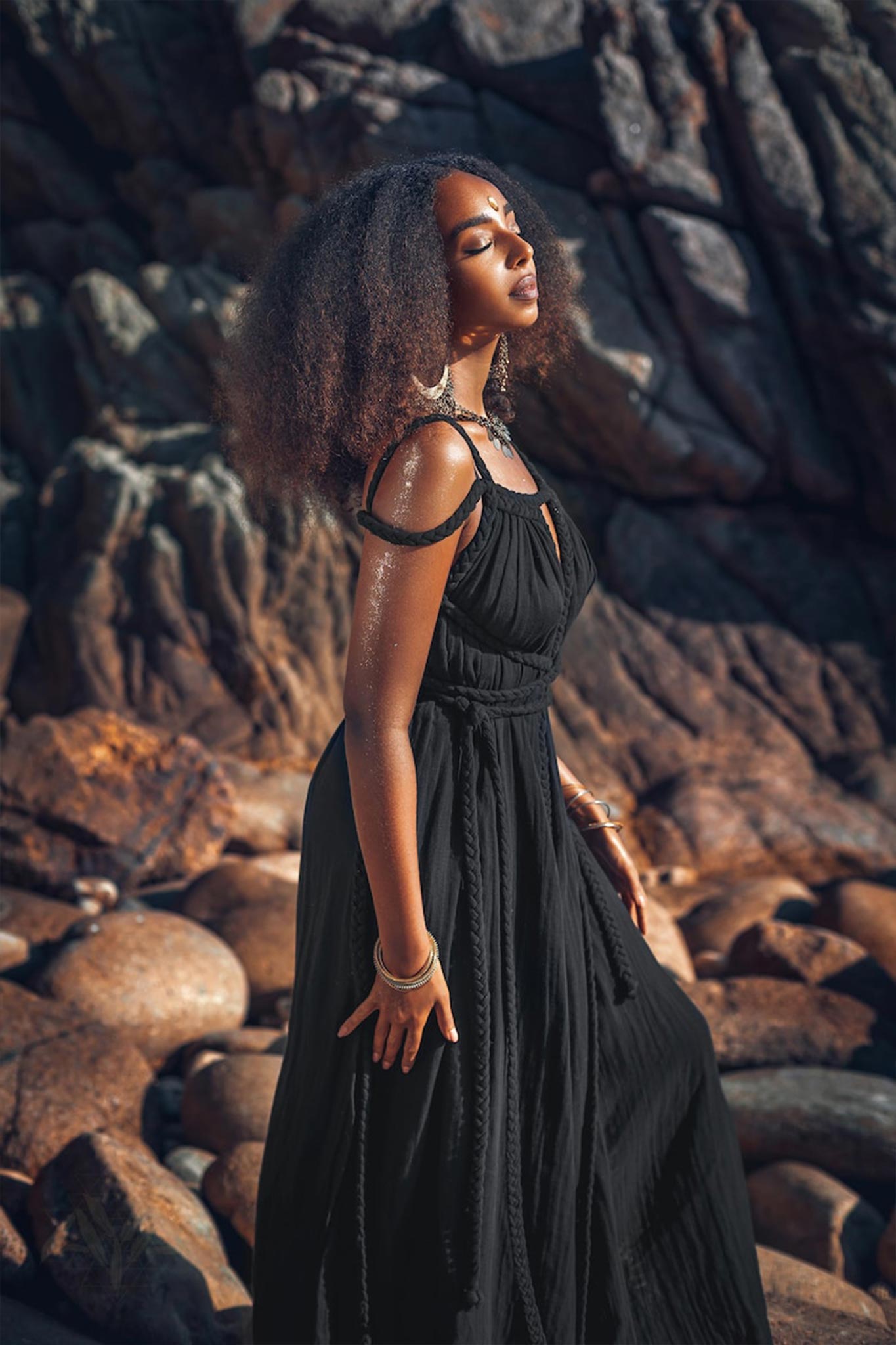 A woman in a Black Greek Goddess Prom Boho Evening Dress by AYA Sacred Wear stands with her eyes closed on a rocky coastline, her curly hair cascading over her shoulders. The sunlight highlights her serene expression and the textures of the rocks surrounding her.